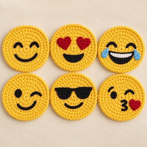 May include: A set of six circular yellow crochet coasters, each featuring a different embroidered emoji face: a smiling face, heart eyes, laughing with tears, a winking face, a face with sunglasses, and a face blowing a kiss with a heart.