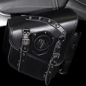 Pode incluir: Alforge de couro preto para motocicleta com detalhes prateados. A bolsa apresenta um emblema decorativo com um design de caveira e é fixada com fivelas e correias. A bolsa é presa a um assento de motocicleta.