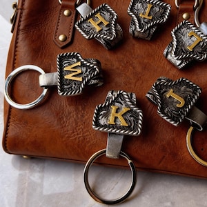 Könnte beinhalten: Silberfarbene Schlüsselanhänger mit goldfarbenen Buchstaben (H, L, T, J, K, W) auf einer braunen Ledertasche. Die Anhänger haben einen dekorativen, gravierten Rand und einen silbernen Schlüsselring.
