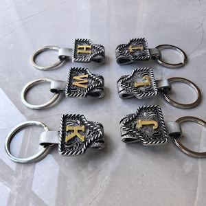 May include: Silver-toned metal keychains with engraved floral patterns and gold-colored initial letters. Each keychain has a silver ring and a leather strap connecting the initial charm. The initials visible are H, I, W, J, K, and J.