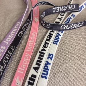 May include: A selection of colourful ribbons with printed text, including "CHANEL", "Happy birthday", and "th Anniversary SUPY 25". The ribbons are in shades of lavender, black, pink, navy, and white. They are displayed against a neutral-toned fabric background.