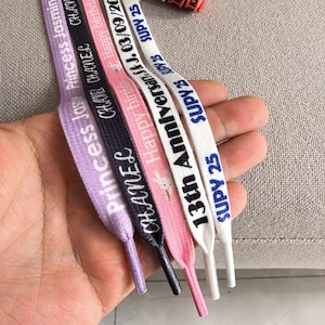 Puede incluir: Una mano sostiene cinco cordones de zapatos de varios colores: lavanda, azul marino, rosa y blanco. Cada cordón presenta texto impreso, incluyendo "Princess Jasmine", "CHANEL", "Happy Birthday", "13th Anniversary" y "SUPY 25". Los cordones son planos y con forma de cinta.