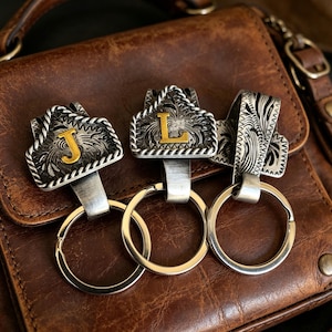 May include: Three silver keychains with decorative, engraved tops. Two keychains feature the letters "J" and "L" in gold. The keyrings are attached to a brown leather surface.