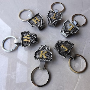 May include: Silver-toned keychains with decorative filigree and gold-colored initial letters. Each keychain has a silver ring and a leather strap. The letters visible are H, L, T, M, K, and J. The keychains are on a light gray surface.