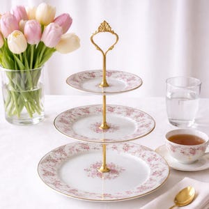 May include: A three-tiered serving stand with floral patterned plates and a gold-colored handle. The plates have a white background with pink floral designs and gold trim. A bouquet of pink and white tulips sits in a glass vase.
