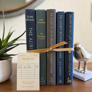 May include: A stack of five hardcover books tied together with a gold ribbon. The spines are dark blue, green, and gray, featuring titles by Tami Hoag, John Grisham, Elizabeth Poliner, M.L. Stedman, and Dana Stabenow. A small cream library card is attached.