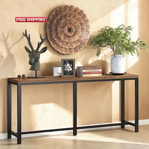 Op de afbeelding: Een lange, rechthoekige consoletafel met een donkerbruin houten blad en zwarte metalen poten. De tafel is versierd met een hertenkopsculptuur, ingelijste kunstwerken, boeken en een vaas met groen. Boven de tafel hangt een grote, ronde houten wanddecoratie.