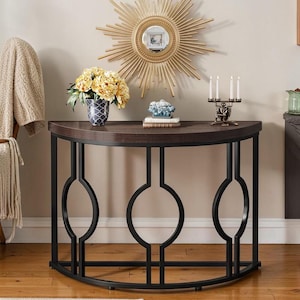 Può includere: Un tavolo consolle a mezzaluna con piano in legno marrone scuro e struttura in metallo nero. La struttura presenta un design decorativo con tre grandi cerchi. Il tavolo è decorato con un vaso di fiori gialli, uno specchio a forma di sole e un candelabro.