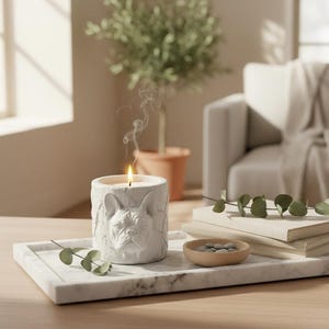 Op de afbeelding: Een witte cilindrische kaarshouder met een gebeeldhouwd gezicht van een Franse Bulldog in reliëf, met een brandende kaars en opstijgende rook. Het staat op een rechthoekig wit marmeren dienblad naast twee boeken, een schaaltje en eucalyptus takjes.