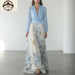 Op de afbeelding: Een lange, vloeiende jurk met een lichtblauwe lijfje en een rok met bloemenprint. Het lijfje heeft een diepe V-hals en lange mouwen. De rok is wit met blauwe en gouden bloempatronen.