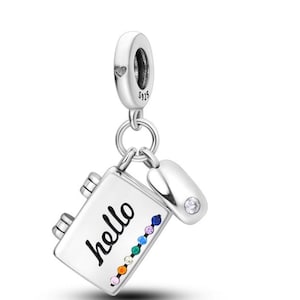 Puede incluir: Un charm plateado con un diseño de cuaderno, la palabra "hello" está escrita en la parte delantera. El charm tiene una serie de gemas de colores a lo largo del borde inferior y un pequeño charm redondo con una sola gema transparente.