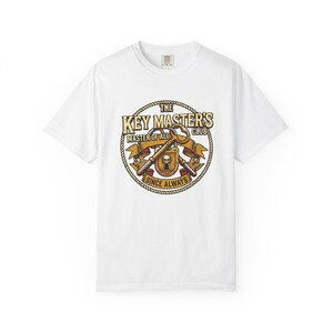 Puede incluir: Camiseta blanca con un diseño circular. El diseño presenta el texto "THE KEY MASTER'S CLUB", "MASTER OF ALL SINCE ALWAYS", llaves, una calavera y un escudo en tonos marrones y dorados.