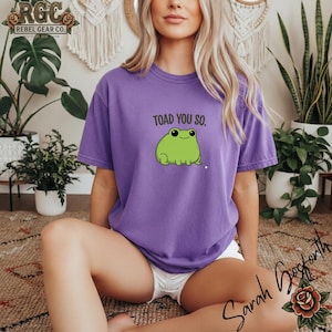 May include: A white ceramic mug with a black handle and the text "But First, Coffee" in a bold, black font. The mug is set against a blurred background. The mug appears to be approximately 10 cm tall.