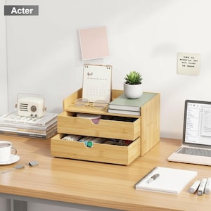 Può includere: Un organizer da scrivania in bambù con due cassetti, un ripiano superiore e una piccola pianta grassa in un vaso bianco. L'organizer contiene un calendario, occhiali e libri. La parola "Acter" è in alto a sinistra.