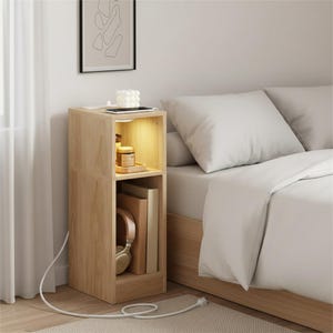 May include: A light-colored wooden nightstand with two open compartments. The top compartment has a small tray with a candle and jar. The bottom compartment holds books and headphones. A white cord and plug are visible.