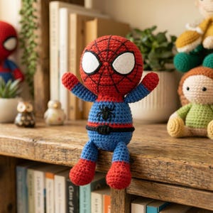 May include: A handmade crochet doll of a superhero character with a red head featuring black web patterns and large white eyes. The body is blue with a black spider emblem on the chest and a black belt. The doll has red arms and legs.
