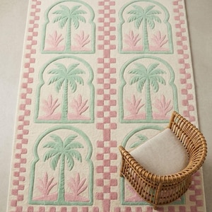 May include: A rectangular rug with a cream background features a repeating pattern of palm trees in arched frames, colored in mint green and pink. The rug is bordered with a pink checkerboard pattern. A wicker chair with a cream cushion sits on the rug.