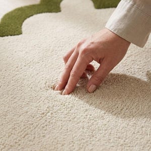 Puede incluir: Primer plano de una alfombra crema texturizada con un dise&ntilde;o en forma de tr&eacute;bol verde en la esquina superior izquierda. Una mano toca la superficie suave de la alfombra, resaltando su textura afelpada.