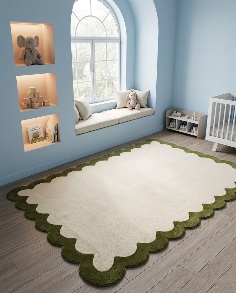Puede incluir: Una alfombra rectangular con un centro color crema y un borde festoneado verde oscuro. La alfombra est&aacute; colocada sobre un suelo de madera clara en una habitaci&oacute;n con paredes azules, un asiento junto a la ventana y una cuna, lo que sugiere una habitaci&oacute;n infantil.