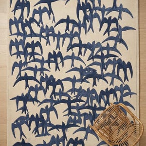 May include: A rectangular rug with a cream-colored background and a pattern of navy blue birds in flight. A small, woven wicker chair is in the lower right corner. The rug is a decorative home accent.
