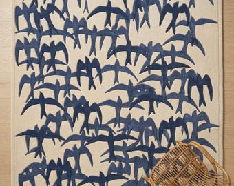 May include: A rectangular rug with a cream-colored background and a pattern of navy blue birds in flight. A small, woven wicker chair is in the lower right corner. The rug is a decorative home accent.