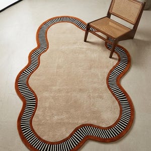 Könnte beinhalten: Ein unregelmäßig geformter Teppich mit organischen Konturen und einer beigen Mitte. Der Teppich ist von einem dicken, rostorangen Rand umgeben, gefolgt von einem schwarz-weiß gestreiften Muster, das dem welligen Verlauf der Außenkante folgt.