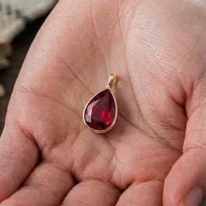 Pode incluir: Um pingente em forma de gota com uma pedra preciosa facetada de cor vermelho profundo, engastada numa moldura de metal dourado polido. O pingente inclui uma pequena argola dourada integrada no topo para fixação a uma corrente, exibindo um tom vermelho claro e vibrante.