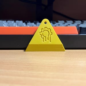 Könnte beinhalten: Ein gelb-goldener, dreieckiger Keycap-Abzieher mit einem Zahnrad- und Tropfendesign. Der Keycap-Abzieher hat oben ein Loch zum Aufhängen. Im Hintergrund befindet sich eine schwarze Tastatur mit orangefarbenen Tasten.