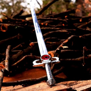 Puede incluir: Una espada plateada con un emblema rojo en la empuñadura, apoyada sobre una superficie de madera. La hoja es larga y reflectante, con un mango detallado. El fondo presenta una pila de troncos de madera, creando una estética rústica.