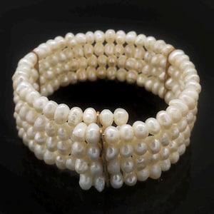 Puede incluir: Un brazalete ancho compuesto por múltiples hilos de perlas blancas cremosas. Las perlas están ensartadas, creando una banda flexible. El brazalete se asegura con un cierre dorado y se muestra sobre un fondo oscuro.