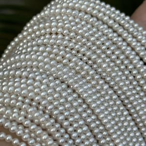 Puede incluir: Primer plano de múltiples hilos de pequeñas perlas blancas y redondas. Las perlas están fuertemente ensartadas, creando una superficie densa y texturizada. Las perlas tienen un aspecto luminoso y brillante, reflejando la luz.