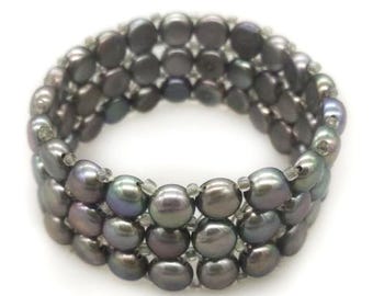 Könnte beinhalten: Ein mehrreihiges Armband bestehend aus drei Reihen dunkelgrauer, runder und glänzender Perlen. Die Perlen sind mit kleinen, klaren, transparenten Zwischenperlen aufgereiht, was ein geschichtetes, kreisförmiges Design mit glatter Oberfläche ergibt.