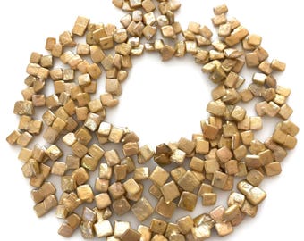 Könnte beinhalten: Eine Ansammlung zahlreicher kleiner, quadratischer Perlen mit metallisch goldener Oberfläche und einer leicht unregelmäßigen, texturierten Struktur. Die Perlen sind in einer kreisförmigen Formation mit offener Mitte angeordnet.
