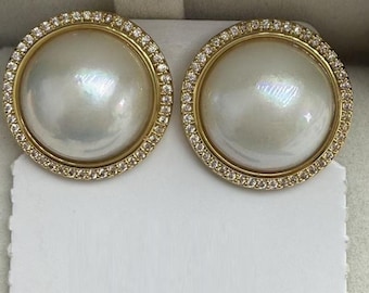 Könnte beinhalten: Ein Paar runde Ohrstecker mit großen, schillernden weißen Cabochon-Mittelstücken. Jeder Ohrring ist von einem dünnen, goldfarbenen Metallrand umgeben, der mit einer Reihe kleiner, klarer, funkelnder Steine besetzt ist, die das Licht einfangen.