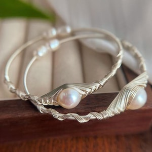 Puede incluir: Dos pulseras de alambre plateado, cada una con una perla grande, redonda y blanquecina. El alambre está intrincadamente tejido alrededor de las perlas y la banda de la pulsera. Las pulseras se exhiben sobre una superficie de madera, sugiriendo un tema de joyería.