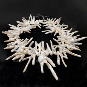 May include: A white, organic-shaped pearl necklace. The necklace is composed of numerous irregular, elongated pearls, strung together to form a circular design. The pearls have a lustrous, iridescent sheen, and the background is black.