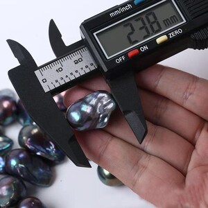 以下が含まれることがあります： 黒いデジタルノギスが、暗く虹色のバロックパールを測定しています。 パールは手で持たれており、ノギスは23.8mmの測定値を表示しています。 他のパールが背景に見えます。