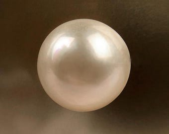 Könnte beinhalten: Eine einzelne, kugelförmige, cremeweiße Perle mit einer glatten, glänzenden Oberfläche. Das Objekt weist einen subtilen irisierenden Schimmer auf, der das Licht auf seiner abgerundeten Form reflektiert und helle Glanzlichter sowie weiche Schatten erzeugt.