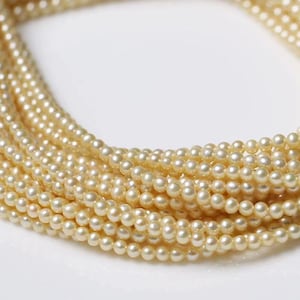 Puede incluir: Primer plano de múltiples hilos de pequeñas perlas de color crema. Las perlas son redondas y de tamaño uniforme, creando una apariencia delicada y elegante. Los hilos están superpuestos, lo que sugiere un collar o una joya similar.
