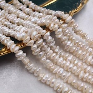 May include: Strands of creamy white, irregularly shaped freshwater pearls. The pearls have a natural luster and are strung together, creating a flowing, organic look. The pearls are displayed on a gold-trimmed surface.