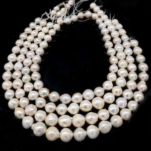 May include: Several strands of luminous, off-white pearls against a black background. The pearls are irregularly shaped, with a smooth, lustrous surface. The strands are arranged in a layered, cascading pattern, showcasing the natural beauty of the pearls.