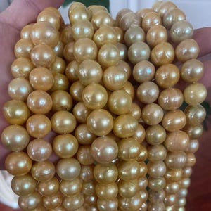 Puede incluir: Primer plano de un conjunto de perlas doradas, redondas y brillantes. Las perlas presentan matices de color que van del oro claro a un tono más profundo. Parecen estar ensartadas, lo que sugiere un collar u otra joya similar.