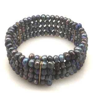 May include: A multi-strand bracelet made of dark, iridescent pearls. The pearls are strung together, creating a flexible design. The bracelet is held together with gold-coloured metal accents. The pearls display hues of blue, purple, and black.