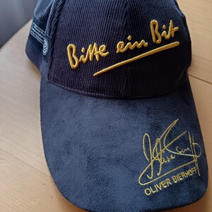 Puede incluir: Gorra de béisbol azul marino con textura de pana. La gorra presenta texto bordado en oro que dice "Bitte ein Bit" y una firma con el nombre "OLIVER BIERHOFF".