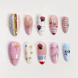 May include: A set of ten almond-shaped artificial nails with various designs. The nails feature stripes, ladybugs, stars, and swirls, with accents of rhinestones and gold detailing. Colors include pink, blue, white, and red.