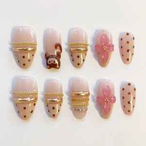 May include: A set of ten press-on nails with a nude base color. Designs include brown polka dots, gold metallic bands, small gold beads, a brown monkey character, and a three-dimensional pink flower accent with a gold center bead on each nail.