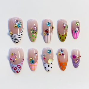 Op de afbeelding: Een set van tien kunstnagels met een nude basis en diverse gekleurde tips, waaronder zebraprint, stippen, strepen en ruitjes. Elke nagel is versierd met kleine metalen kraaltjes en veelkleurige bedels in de vorm van edelstenen.