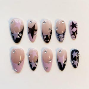 Puede incluir: Un juego de diez uñas postizas en forma de almendra con base nude y patrones de estrellas en negro y morado claro. Varias uñas incluyen puntas francesas negras, texturas de piel de serpiente y pequeños diamantes transparentes aplicados.
