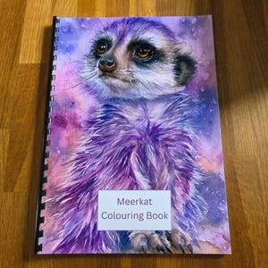 May include: A spiral-bound colouring book featuring a watercolour illustration of a meerkat on the cover. The meerkat is rendered in shades of purple and blue against a vibrant, cosmic-style background. The text Meerkat Colouring Book is printed.