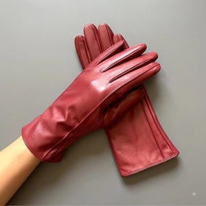 May include: A pair of deep red leather gloves featuring a smooth finish, stitched detailing along the fingers, and a clean, straight-cut wrist opening. The gloves are layered over one another, showcasing the rich, saturated colour and texture.